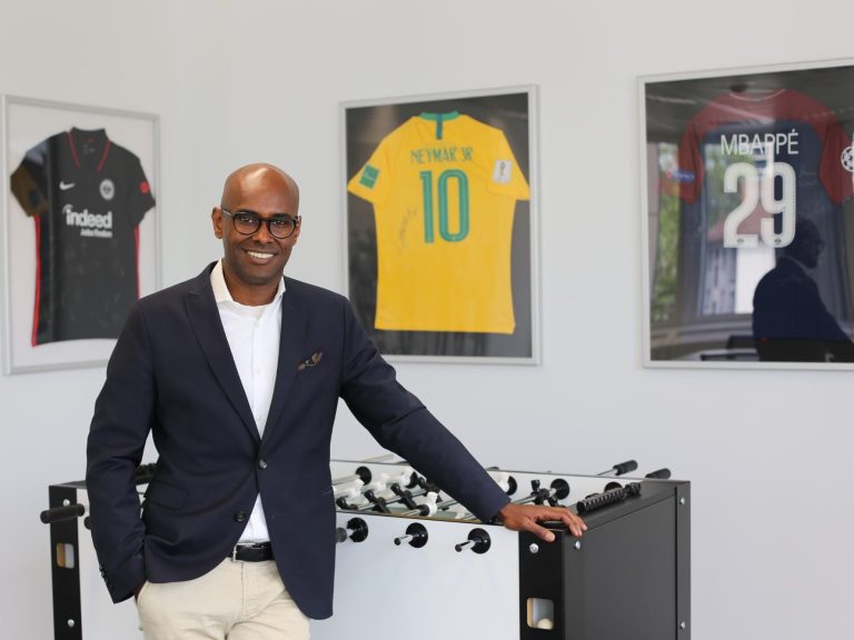 Gregor Ghirmay steht neben dem Büro Tischkickertisch und vor den Fußballtrikots von Mpappe, Neymar und Eintracht Frankfurt